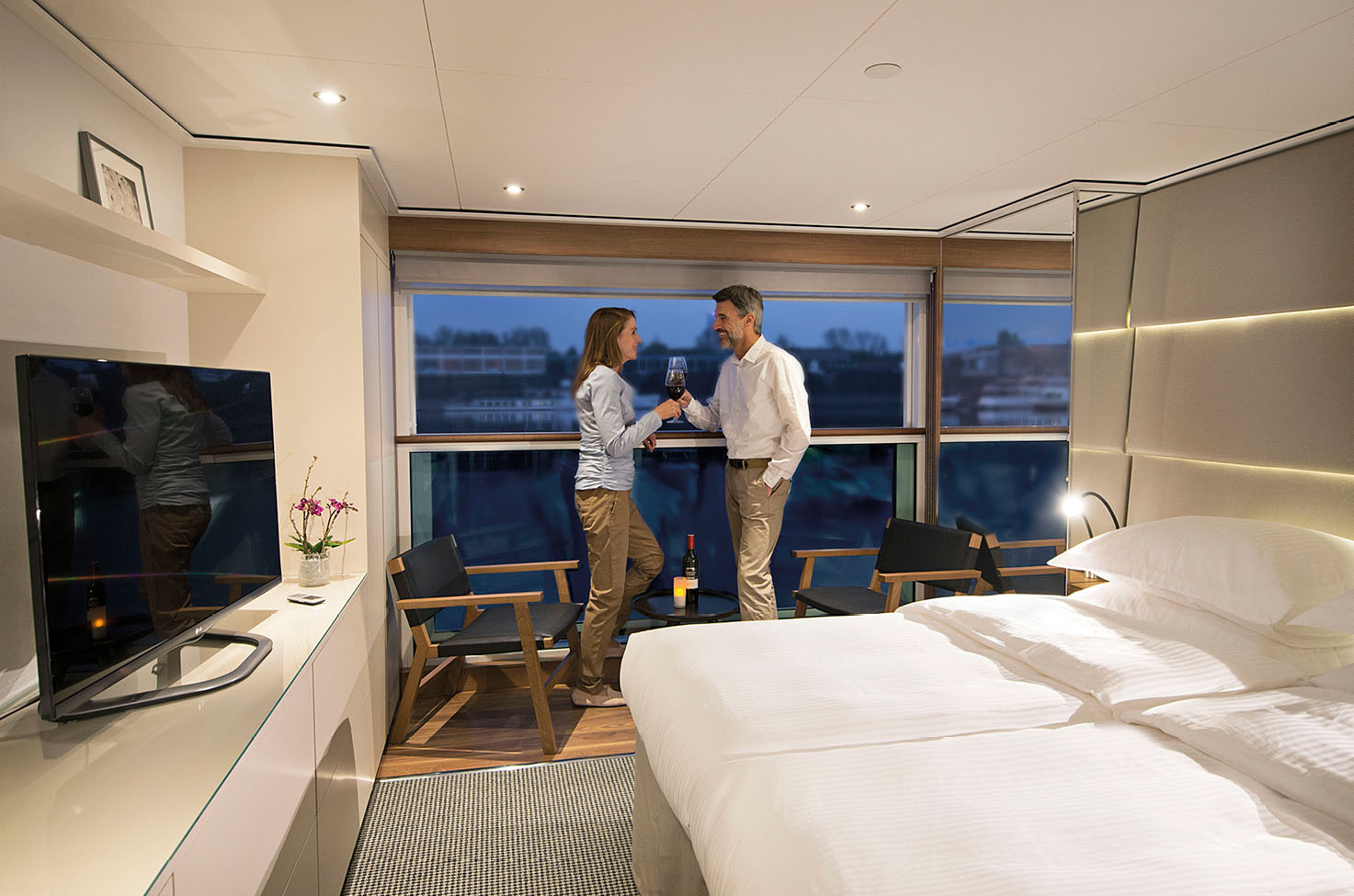 Man and woman in their spacious cabin, standing near the window at nighttime, close to the white-linen bed