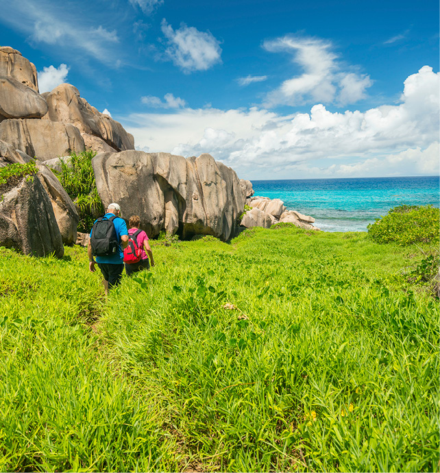Two people walking through the green grass with turquoise waters and the blue sky as a backdrop on La Digue Island. 