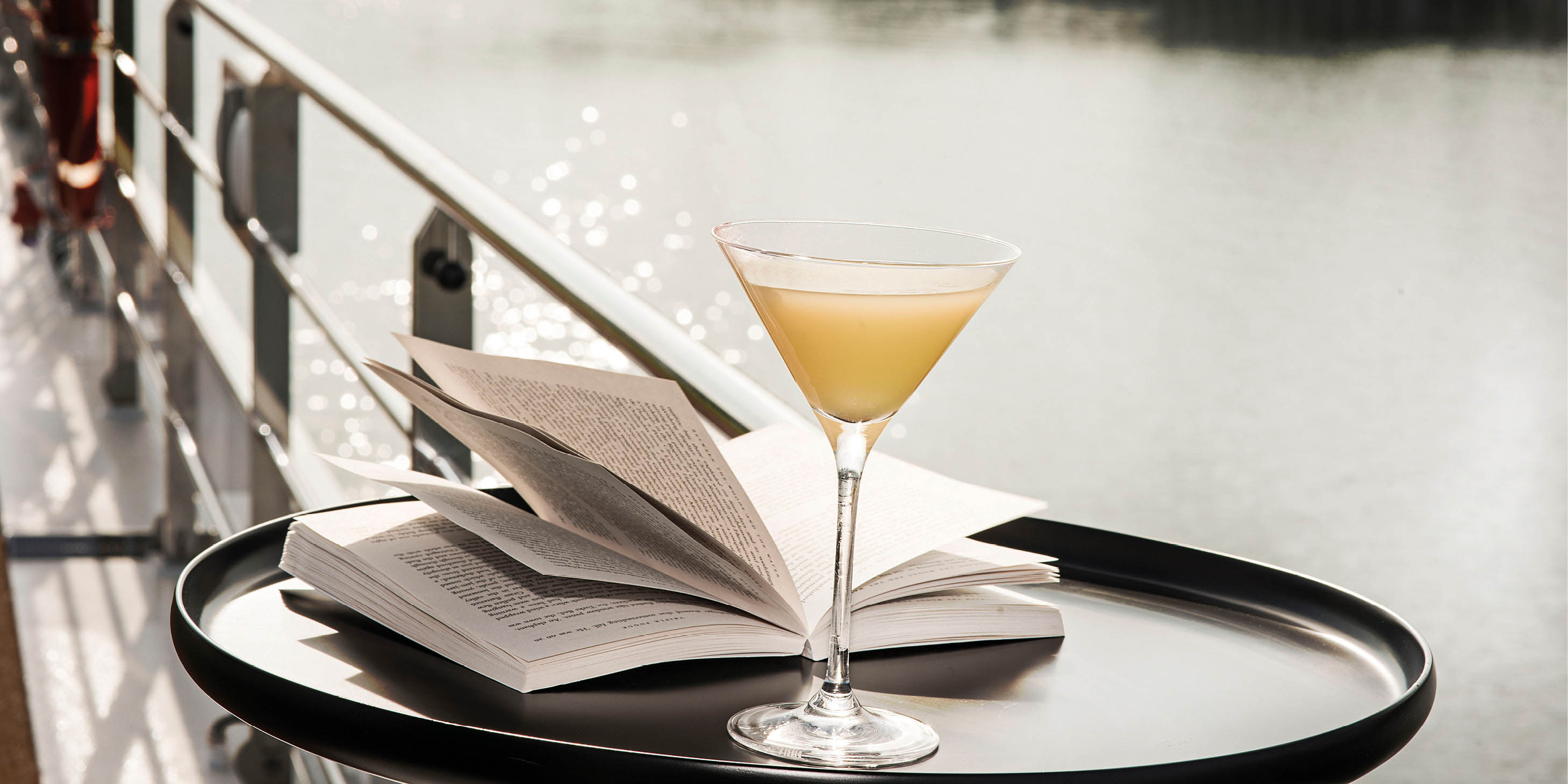 An open book next to a cocktail on a table on a cruise deck, overlooking a river, with a cruise ship in the background