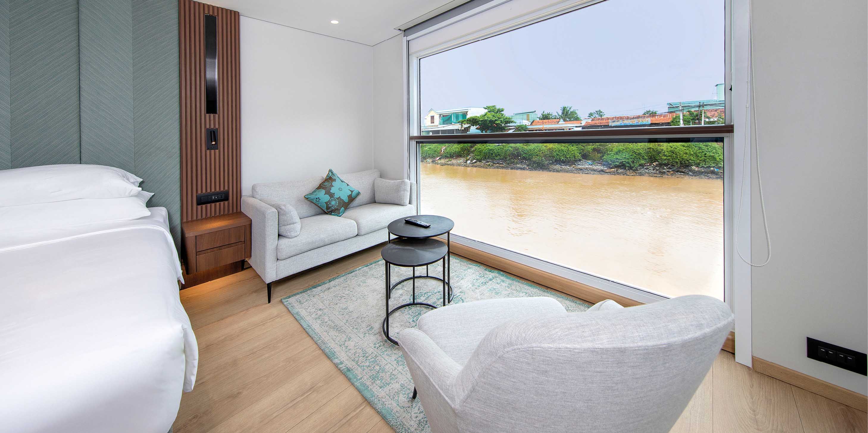 A two-seater sofa and chair next to a large balcony with views of the Mekong River, with a crisp, white bed to the side of the room