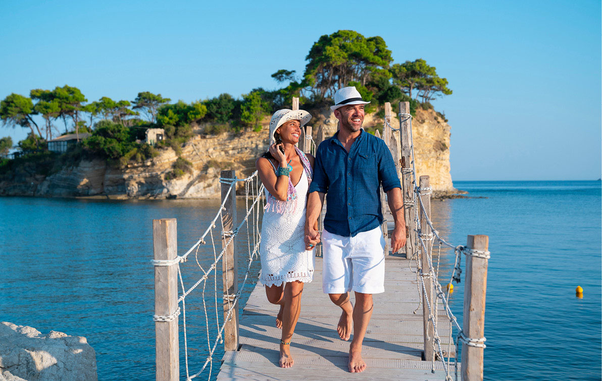 Couple holding hands and smiling as they walk along a bridge over the blue sea from a cliffy, green island.